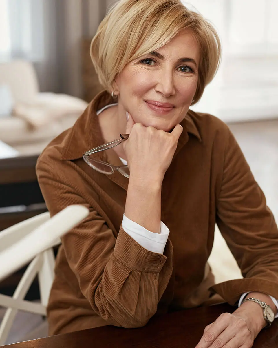 mature woman wearing brown coat with her glasses in her hand